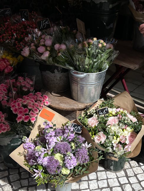 A variety of fresh flower bouquets displayed on a wooden surface, featuring soft pink roses, lavender-purple lisianthus, and white and pink anemones, all wrapped in brown craft paper or stored in meta