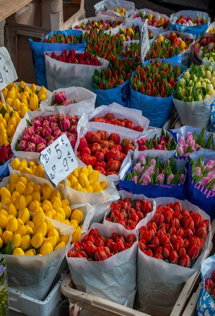 A vibrant display of fresh tulips arranged in large paper and plastic wrapping bags at a flower market, featuring a variety of colours including yellow, pink, orange, red, purple, and green buds. The 