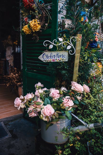 A floral display outside a garden centre featuring a pastel pink rose bouquet in a silver watering can, surrounded by lush green foliage and potted plants. In the background, there is a green wooden g