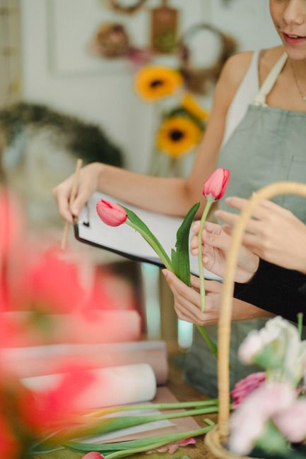 A florist in a workshop holding a bouquet of two pink tulips with green stems and leaves, examining or arranging them. The flowers are fresh with smooth, vibrant petals, and the bouquet is partially w