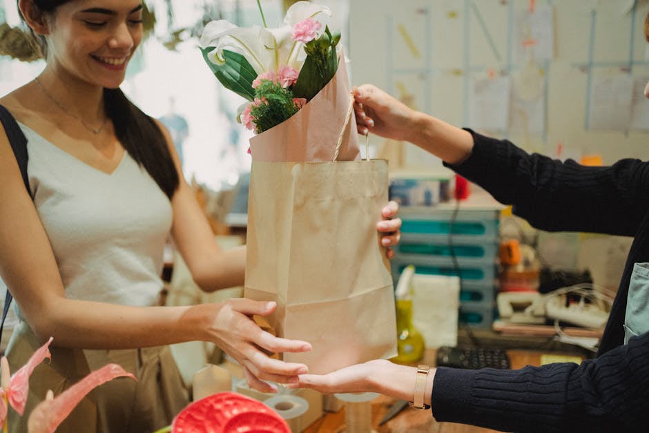A woman with long dark hair, dressed in a white sleeveless top, is smiling as she receives a bouquet of fresh flowers from a florist. The bouquet, wrapped in pink tissue paper and placed inside a beig