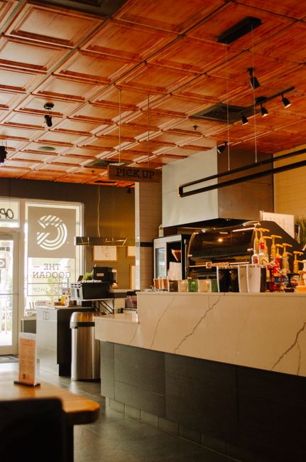 The image depicts the interior of a modern café with a warm wooden coffered ceiling and contemporary lighting fixtures. A white marble counter with subtle veining features a selection of coffee-makin