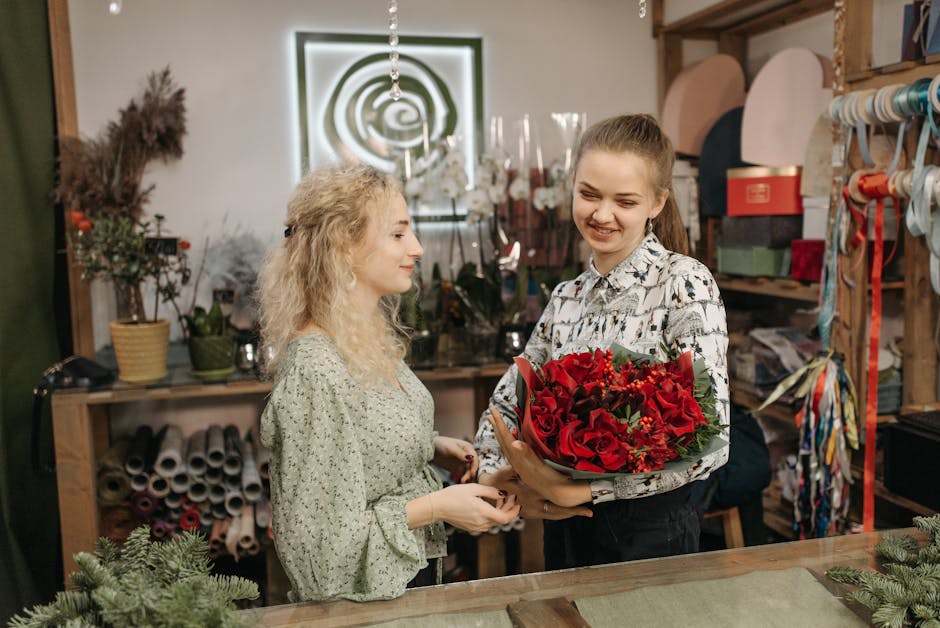 A young woman with light brown hair tied back, wearing a white shirt with black patterns, is handing a bouquet of fresh red roses and greenery wrapped in brown paper to a woman with curly blonde hair 