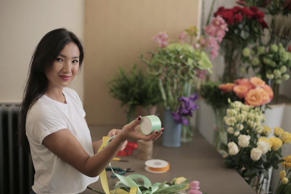 A young woman with long dark hair, wearing a white t-shirt, is smiling and holding a roll of pale green floral tape in her right hand. She is in a well-lit floral workshop with a neutral beige wall in