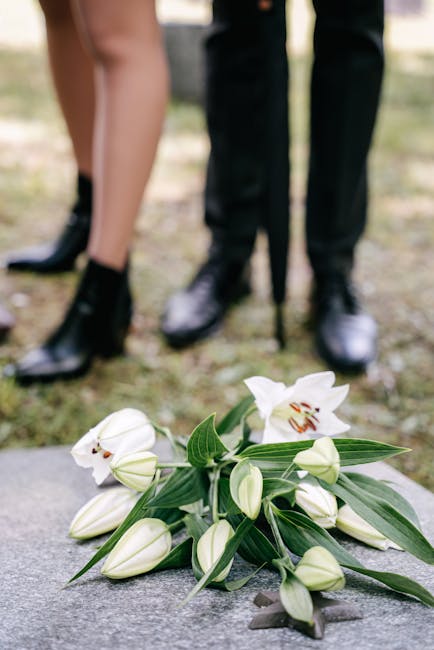 A bouquet of white lilies and unopened lily buds with long green leaves, arranged with some petals and flowers resting on a stone surface. In the background, the lower legs and feet of a person dresse