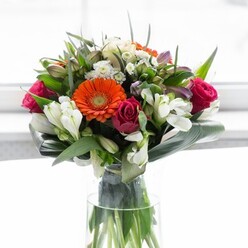 This splendid bouquet unfurls like a celebration of Redhill's local charm and contemporary flair, artfully created for moments of genuine connection. At its centre, glowing orange gerbera daisies share space with gentle cream-peach companions, each highlighted by a dark central eye that draws the gaze. Lively magenta roses cluster among them, their tightly wound petals whispering of special occasions and affectionate wishes delivered right to doorsteps across Redhill. Pure white alstroemeria, joined by dainty sprays of white blossoms, fill the spaces between larger blooms with crisp radiance and textural variety, just as the trees and hedges brighten Redhill's neighborhoods. Gentle blush pinks surface in the ensemble, adding richness and depth, while the structure is defined by glossy, broad aspidistra leaves that wrap around the bouquet's base in a supportive, verdant frame. Smaller, lively green leaves and wisps complete the composition, filling each gap to evoke a garden's natural abundance. The design's upright lines are mirrored in a crystal-clear glass vase, through which the green stems remain visible, offering a fresh, organic touch. Soft natural light floods the scene, causing the vibrant palette to glow gently-suffusing the bouquet with an atmosphere of joy, gratitude, and celebration. Ideal for local gatherings, special gifts, or simply brightening a friend's home, this bouquet stands as a testament to Redhill's skilled florists and the natural beauty found throughout the community.